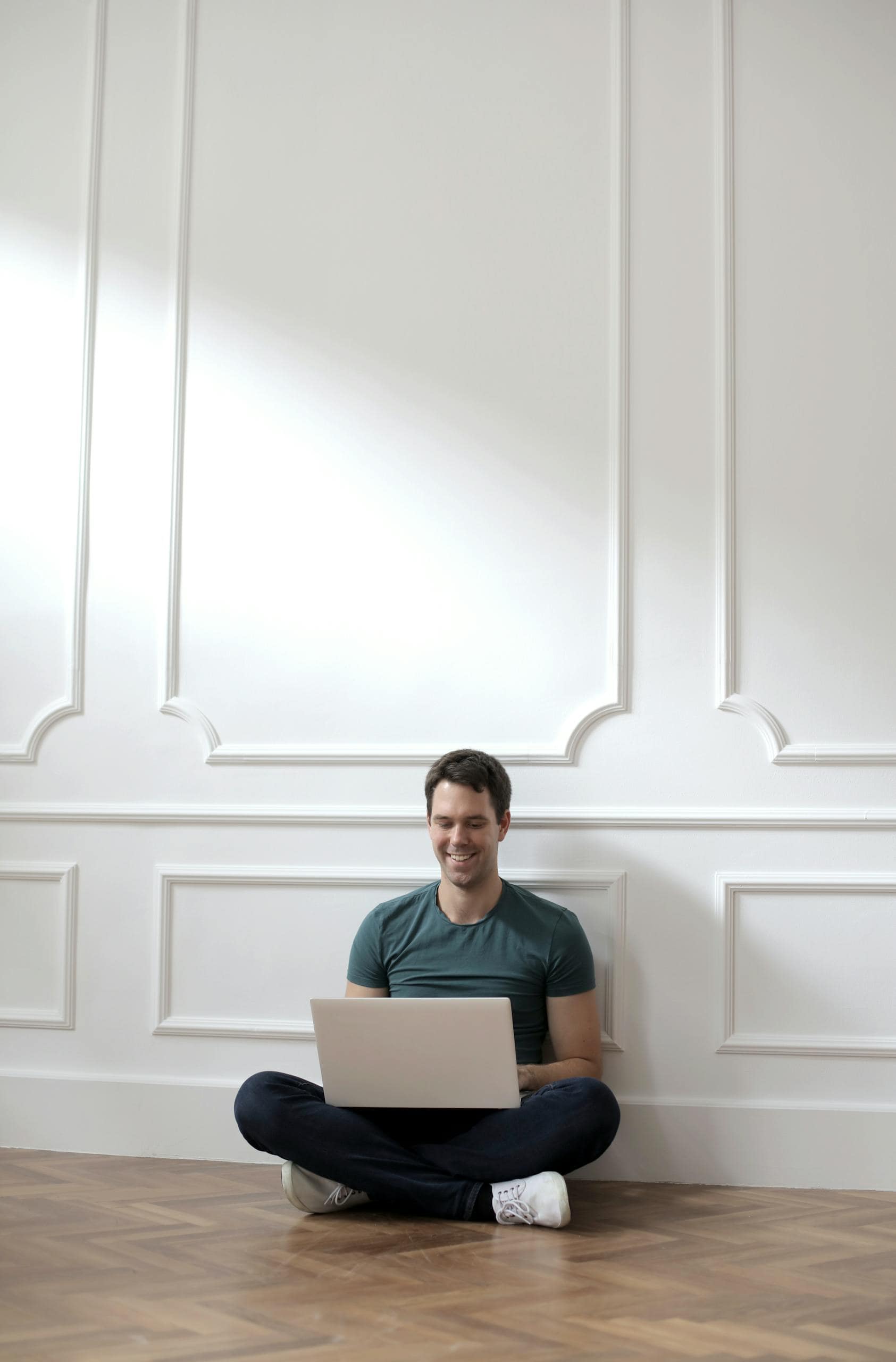Young man smiling while working remotely on a laptop sitting on the floor at home.